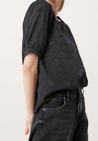 Sort bluse med korte, puffede ærmer, der har et tekstureret design og åben vævning. Parret med mørkegrå jeans, som fremhæver en afslappet stil.