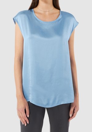 Jasnoniebieska bluzka z krótkim rękawem wykonana z gładkiej tkaniny, z zaokrąglonym dekoltem i zakrzywionym dołem, zestawiona z ciemnymi spodniami.