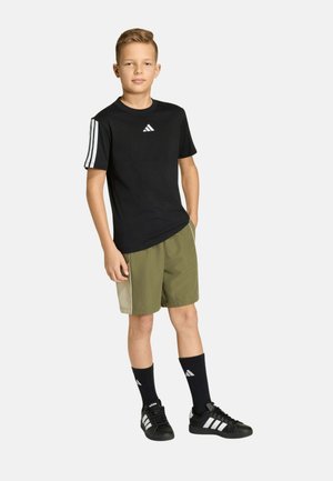 Niño vistiendo una camiseta negra de Adidas, pantalones cortos verdes, calcetines negros de Adidas y zapatillas negras de Adidas, de pie con las manos en los bolsillos.