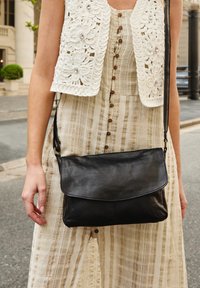 Black leather crossbody bag with a flap closure, smooth texture, and a thin adjustable strap. Worn over a beige striped dress with a lace vest.
