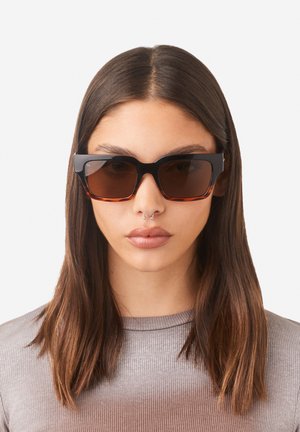 Occhiali da sole neri e tartarugati con montatura squadrata, lenti scure e finitura liscia, indossati da una persona con capelli lunghi e lisci.