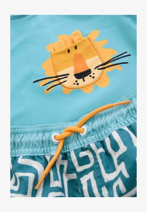 Törölközőnadrág türkiz színben, sárga és barna oroszlánfej hímzéssel. Fehér geometrikus mintákkal rendelkezik, állítható narancssárga zsinórral.