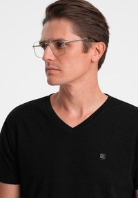Černé tričko s výstřihem do V vyrobené z měkké látky, s malým čtvercovým logem v dolním rohu. Čiré kovové brýle s minimalistickým designem.