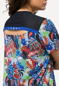 Camisa de manga corta con un vibrante patrón floral en varios colores. La parte trasera cuenta con un yugo negro sólido con ribete azul y puntos decorativos.