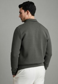 Sweatshirt gris foncé avec un col et des poignets côtelés, design ajusté, texture lisse, associé à un pantalon de couleur claire.