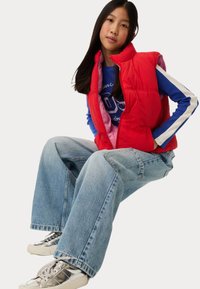 Rode gewatteerde vest over een blauwe shirt met lange mouwen, gecombineerd met lichtblauwe wijde jeans en metallic zilveren sneakers.