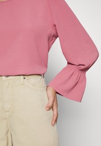 Femme portant un chemisier rose à manches longues avec des manches cloche froncées et un pantalon beige taille haute avec des poches avant visibles.