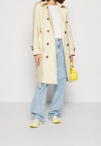 Beige kaksirivisillä napituksilla varustettu trenssitakki, yhdistetty vaaleisiin farkkukapeisiin leveälahkeisiin farkkuihin ja valkoisiin lenkkareihin, joissa on keltaisia yksityiskohtia. Pieni keltainen käsilaukku.