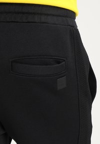 Černé pánské tepláky se strukturovaným bočním panelem, s malou nášivkou s logem a zadní kapsou s obdélníkovým otevřením.