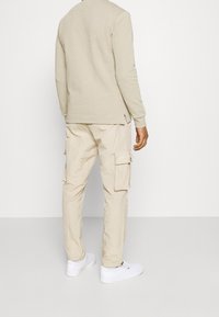 Homme portant une chemise beige à manches longues, un pantalon cargo beige clair avec des poches latérales et des baskets blanches, debout devant un fond blanc uni.