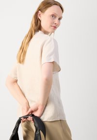Beige lněný top s krátkým rukávem, volnější střih, boční rozparek a přírodní struktura; kombinovaný se světle hnědými kalhotami a držící černou tašku.