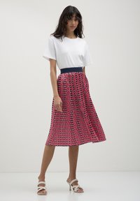 Donna in piedi che indossa una t-shirt bianca a maniche corte, una gonna midi plissè a fantasia rosa e sandali con tacco bianchi su uno sfondo semplice.