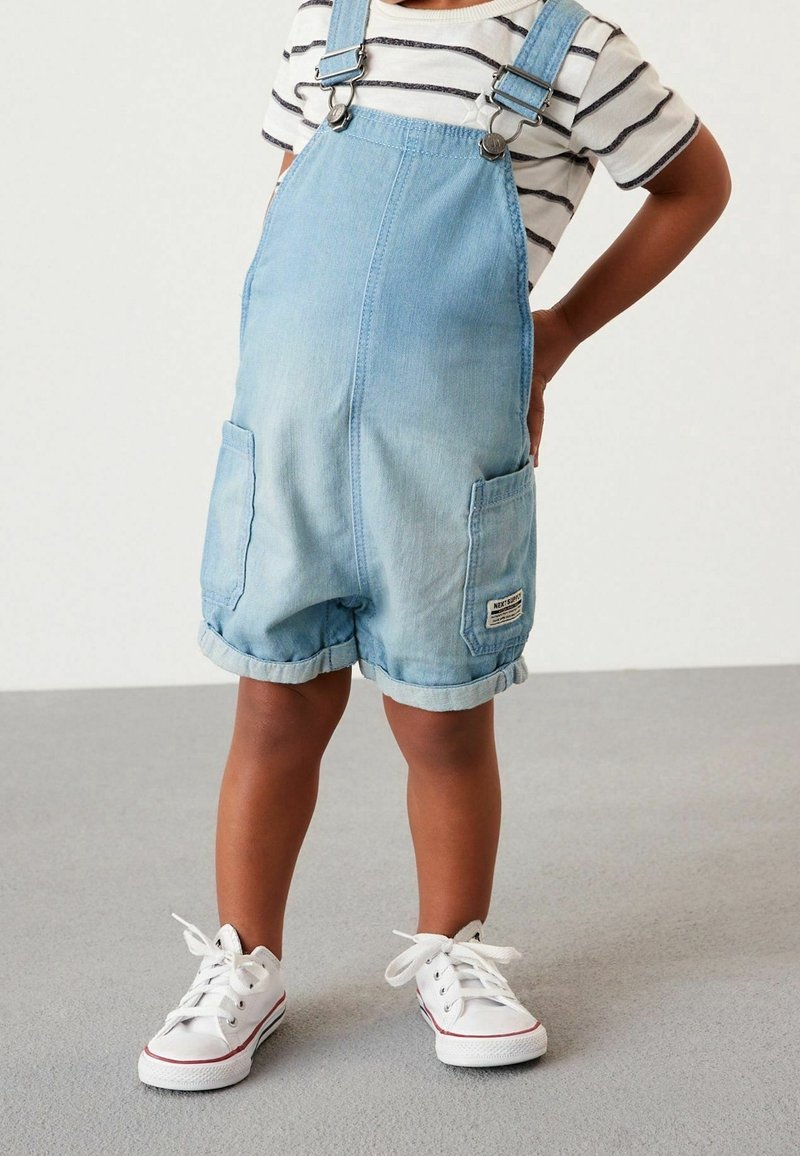 Enfant portant une salopette courte en denim bleu clair, un t-shirt rayé blanc et noir, et des baskets blanches, debout sur un sol gris.