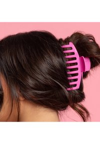 Rosa plastkam fäst i mörkt, vågigt hår som håller en knut på plats. Slät yta och öppen design med breda tänder. Bakgrunden är rosa.