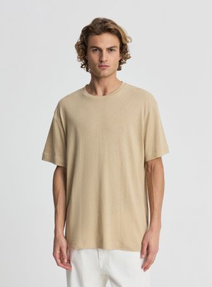 Giovane uomo con capelli ricci che indossa una maglietta beige oversize a maniche corte e pantaloni bianchi, in piedi davanti a uno sfondo chiaro e semplice.
