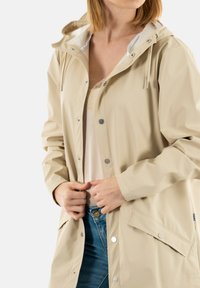 Chaqueta de lluvia impermeable beige con capucha, botones a presión y bolsillos laterales. Presenta una textura suave y cordón ajustable en la capucha.