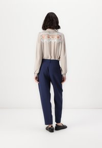 Beige bluse med blonde detaljer på baksiden, kombinert med marineblå bukser. Svarte sko med dekorativt utstyr. Glatt, lett stoff.