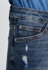 Jeans in denim di colore blu scuro con un motivo usurato, dotati di una piccola tasca frontale, hardware in metallo e un'etichetta con il marchio.