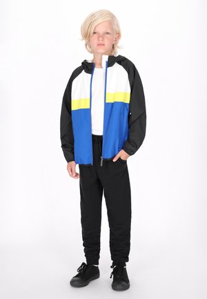 Blauwe, witte en zwarte windjack met een gele streep; met rits; gecombineerd met zwarte broek en zwarte sneakers; gladde stofstructuur.