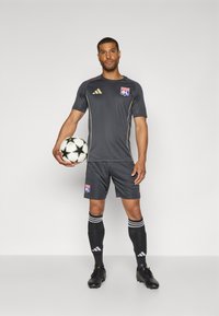 Uniforme de soccer gris avec des accents dorés, arborant un logo sur la poitrine. Le joueur tient un ballon de soccer noir et blanc avec un motif étoilé. Chaussures à crampons noires.