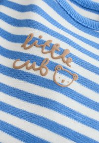 Tkanina plava i bijela prugasta s bež uzdignutim tekstom "mali medvjed" i jednostavnim obrubom lica medvjedića izvezanim na njoj.