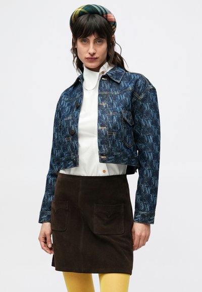 Orezaná džínsová bunda z modrej textúrovanej látky s vzorom, zlatými akcentmi a tmavohnedá semišová sukňa s dvoma vreckami. Žlté pančuchy.
