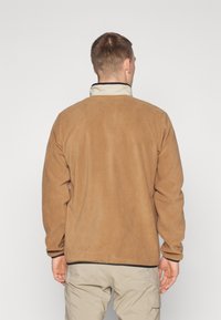 Mand iført tan fleecejakke og beige cargo-bukser, stående med ryggen til mod en ensfarvet lys baggrund.