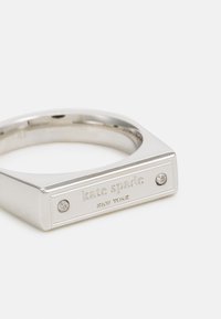 Inel din metal de culoare argintie, având un vârf dreptunghiular plat, pe care este gravat "kate spade NEW YORK" și două pietre clare încorporate.