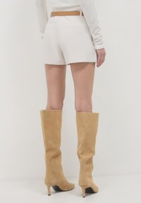 Weiße Shorts mit hoher Taille und einem beigen Gürtel, kombiniert mit knielangen beigen Wildlederstiefeln und einem strukturierten Langarmoberteil.