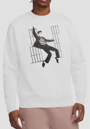 Person trägt weißen Pullover mit schwarz-weißem Motiv eines tanzenden Mannes vor vergitterten Fenstern.