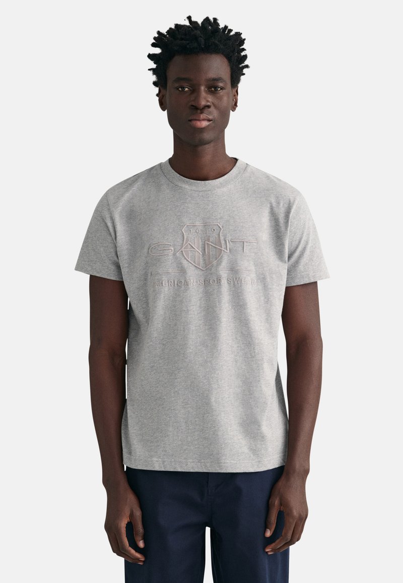 T-shirt en coton gris à manches courtes, présentant un design de logo en relief de couleur gris plus foncé. Encolure ronde, coupe décontractée et texture unie.