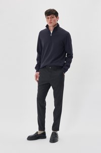 Jūrų laivyno zip-up puloveris, šviesiai dryžuotos juodos kelnės, struktūrinė medžiaga, juodi loafers su masyviu padu. Modelis stovi prieš paprastą foną.