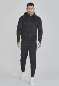 Crna hoodie s kengurujem i toniranim logotipom, u uskom kroju. Uparen s crnim jogger hlačama i bijelim tenisicama.
