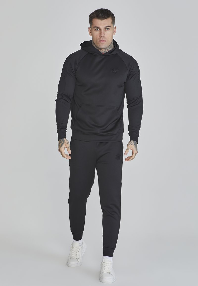 Crna hoodie s kengurujem i toniranim logotipom, u uskom kroju. Uparen s crnim jogger hlačama i bijelim tenisicama.