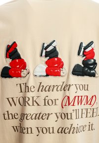 Camiseta beige que presenta tres figuras de dibujos animados bordadas al revés, vistiendo ropa roja, negra y blanca. Texto impreso debajo en una fuente en negrita.