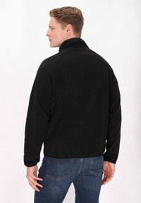 Giacca di pile nera con colletto alto, maniche lunghe e texture liscia, abbinata a jeans di denim blu. Posizionata da un angolo posteriore.