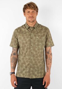 Chemise à manches courtes, de couleur olive, à boutons, avec un motif répétitif de feuilles de palmier, dotée d'une poche poitrine et de boutons marron.