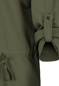 Olijfgroene kledingstof van dichtbij met een geplooide mouwontwerp, knoopdetails en een trekkoord in de taille voor aanpassing.