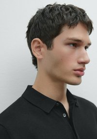 Polo shirt nera con colletto classico, caratterizzata da dettagli con bottoni. Volto del modello di profilo, con capelli corti e texturizzati e sfondo neutro.