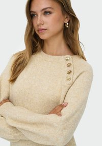 Beige trui van zachte brei-stof, met een ronde halslijn en gouden knoopaccenten op de linkerschouder. Gefacetteerd en gezellige ontwerp.