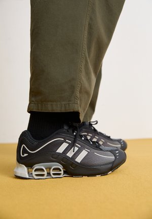 Black and gray athletic shoes with white detailing worn with black socks and olive green pants standing on a yellow surface.