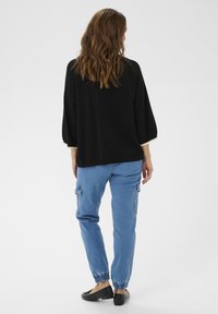 Barna hull�ámos hajú nő, aki laza fekete pulcsit, kék farmer cargo nadrágot elasztikus mandzsettával és fekete lapos cipőt visel, háttal áll.