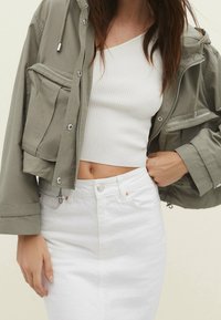 Vrouw in een cropped witte geribbelde top, een hoge witte rok en een losse olijfgroene jas met grote ritspockets.