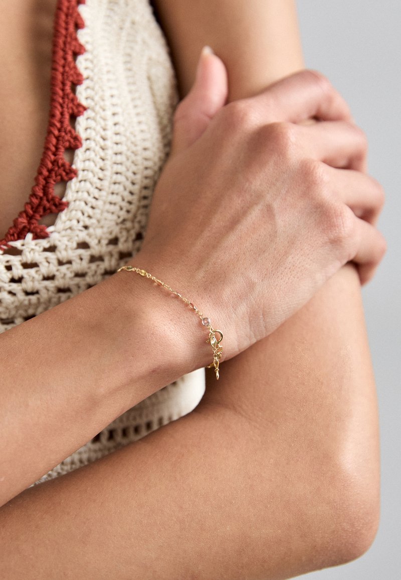 Kuldne kett-randmete, millel on väikesed helmed ja dekoratiivne ripats, kantud randmel. Taustal on tekstuuriga heledat värvi särk.