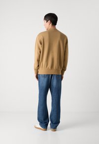 Sudadera marrón con puños y dobladillo de canalé, combinada con jeans holgados de color azul. El modelo se encuentra de pie frente a un fondo blanco liso.