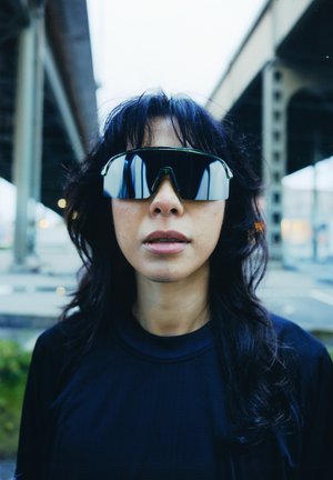 Czarne, lustrzane okulary przeciwsłoneczne o sportowym designie, o dużych, kanciastych kształtach. Noszone przez osobę o ciemnych włosach, na tle miejskim.