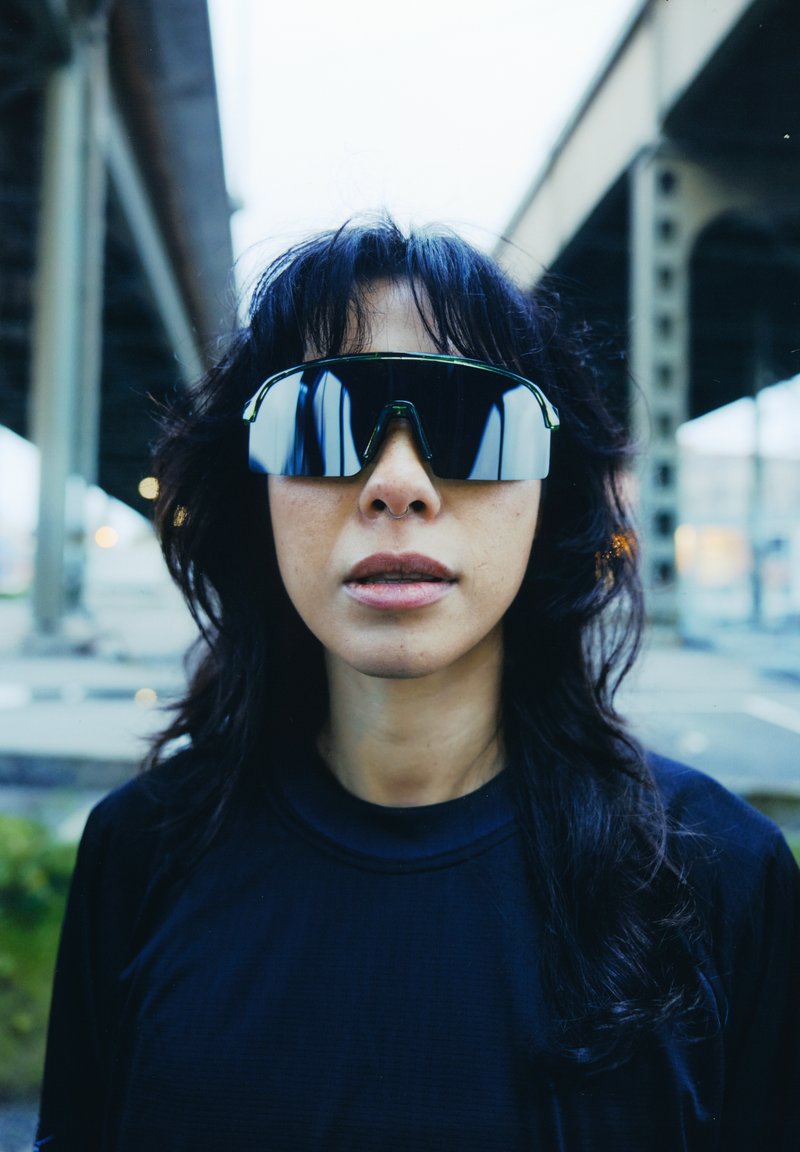 Čierne reflexné slnečné okuliare so športovým dizajnom, nadrozmerné a uhlové tvary. Nosené osobou s tmavými vlasmi, na mestskom pozadí.