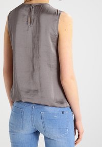 Vrouw in een mouwloze grijze top met een knoopsluiting en een sleuf aan de achterkant, gedragen met lichtblauwe spijkerbroek, gezien van achteren.