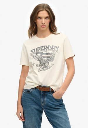 Kremowa bawełniana koszulka z grafiką przedstawiającą orła oraz napisem "SUPERDRY" w kolorze czarnym. Krótkie rękawy i luźny krój, zestawiona z niebieskimi dżinsami.