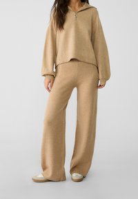 Jasnobrązowy, miękki sweter z kapturem z ćwierć zamkiem i ściągaczami, zestawiony z dopasowanymi spodniami z szerokimi nogawkami. Swobodny i komfortowy wygląd.
